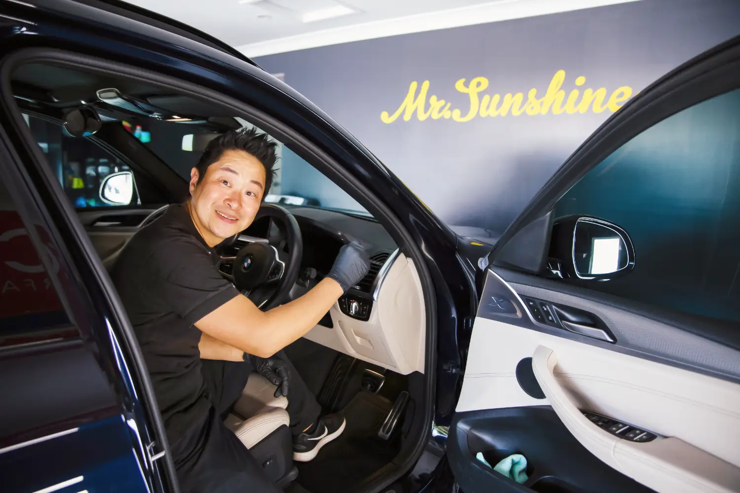 A person seated in the driver's seat of a car, smiling at the camera. The vehicle door is open, and the words "Mr. Sunshine" are visible on a wall in the background.