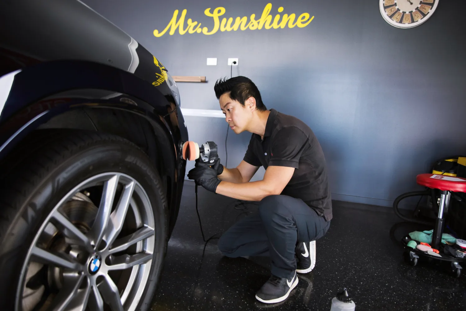 A person is polishing a black car's fender using a handheld buffer in a garage, with "Mr. Sunshine" written on the wall in the background.