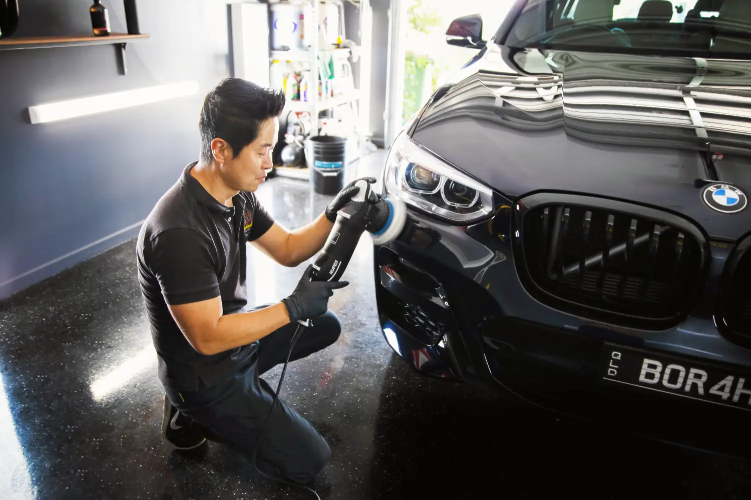 A person kneels while using a polishing machine on the front of a black BMW car in a garage.