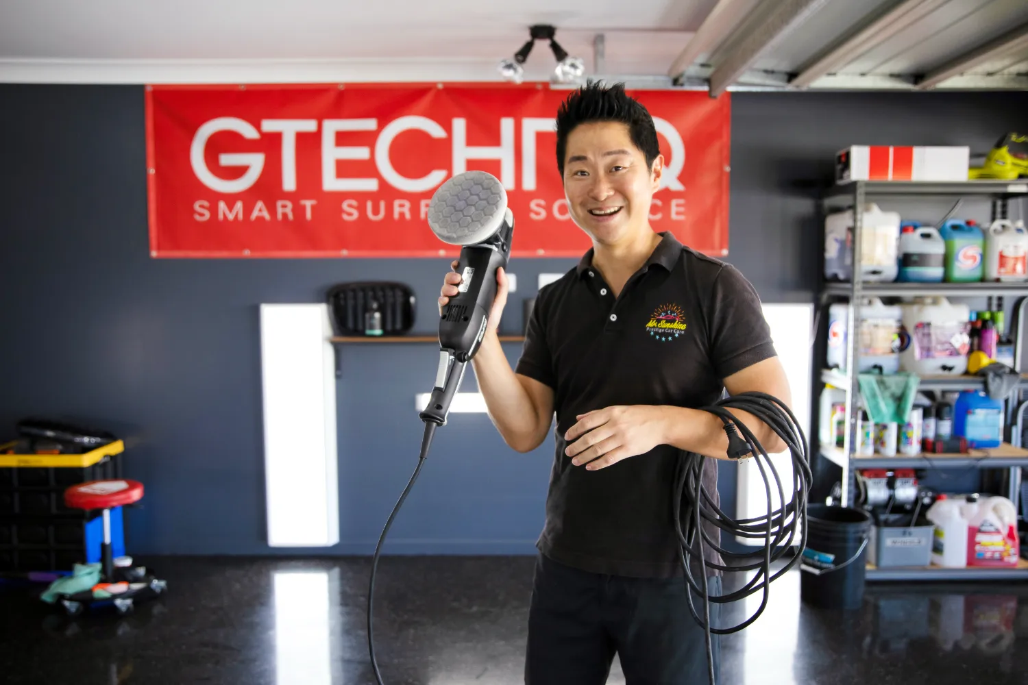 A person stands in a garage holding a power tool next to a red Gtechniq banner. Shelves filled with various items are in the background.