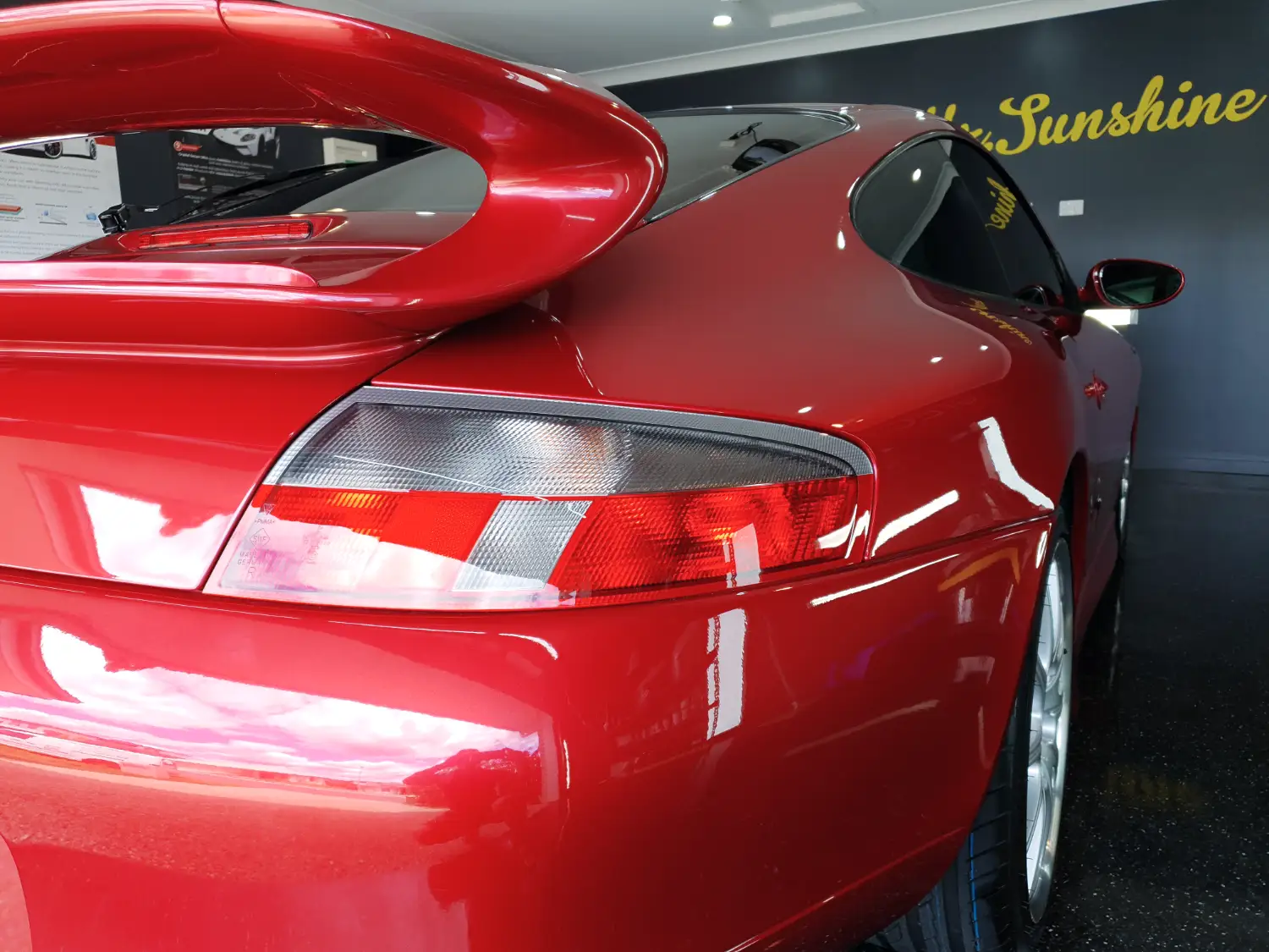 Close-up of the rear end of a red sports car with a large rear spoiler, parked indoors near a wall with the words "sandy sunshine" in the background.