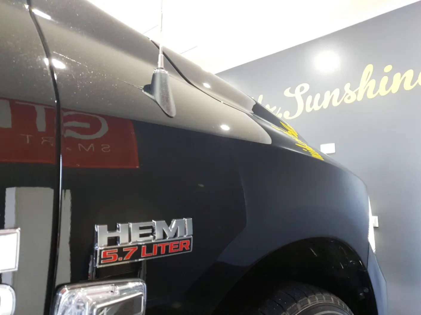 Close-up of a black vehicle with a "HEMI 5.7 Liter" badge on the side and partial view of a headlight and tire. Script lettering reading "Sunshine" is visible in the background.