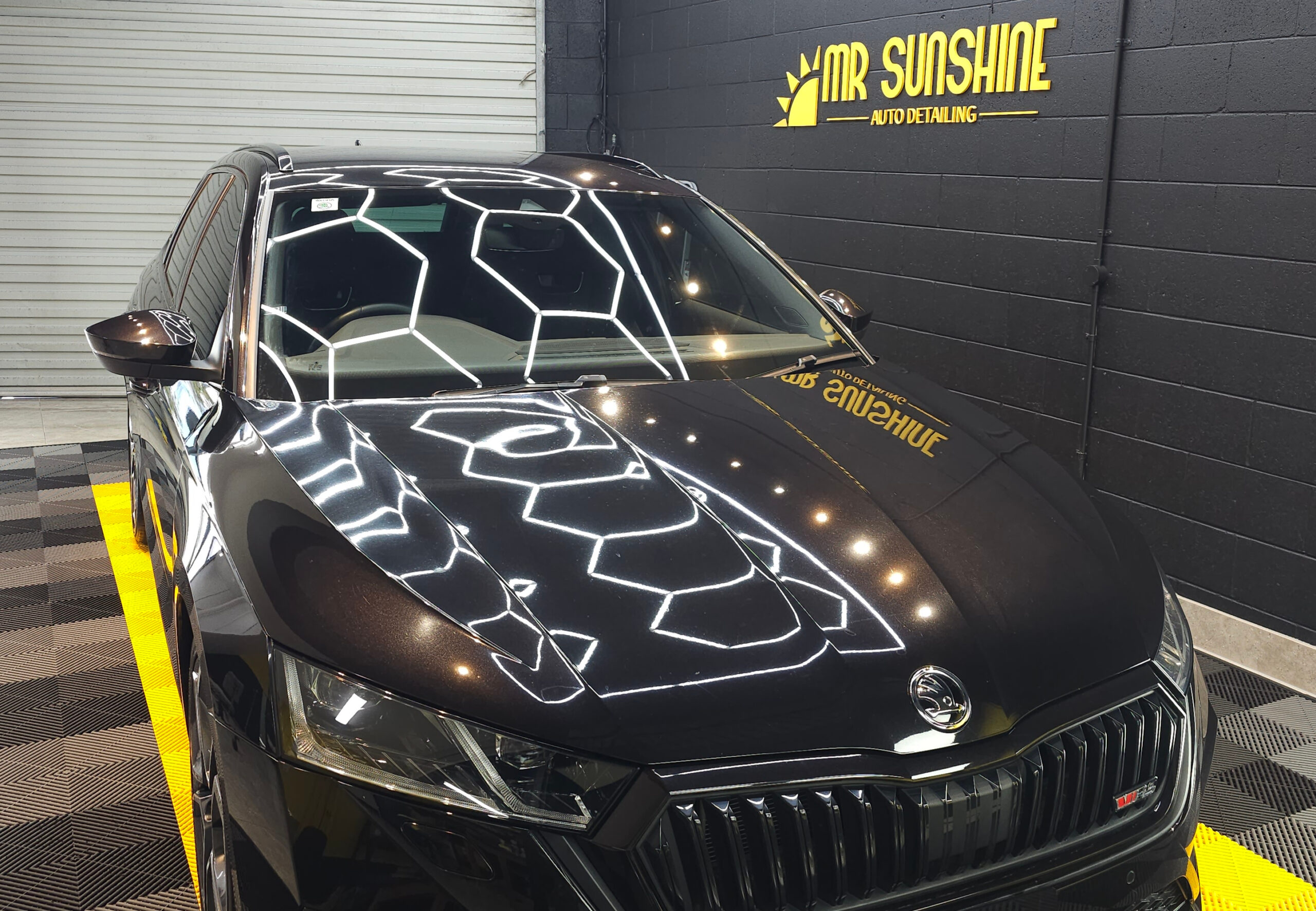 A black Skoda car is parked indoors at Mr Sunshine Auto Detailing, with geometric ceiling lights reflected on its hood.