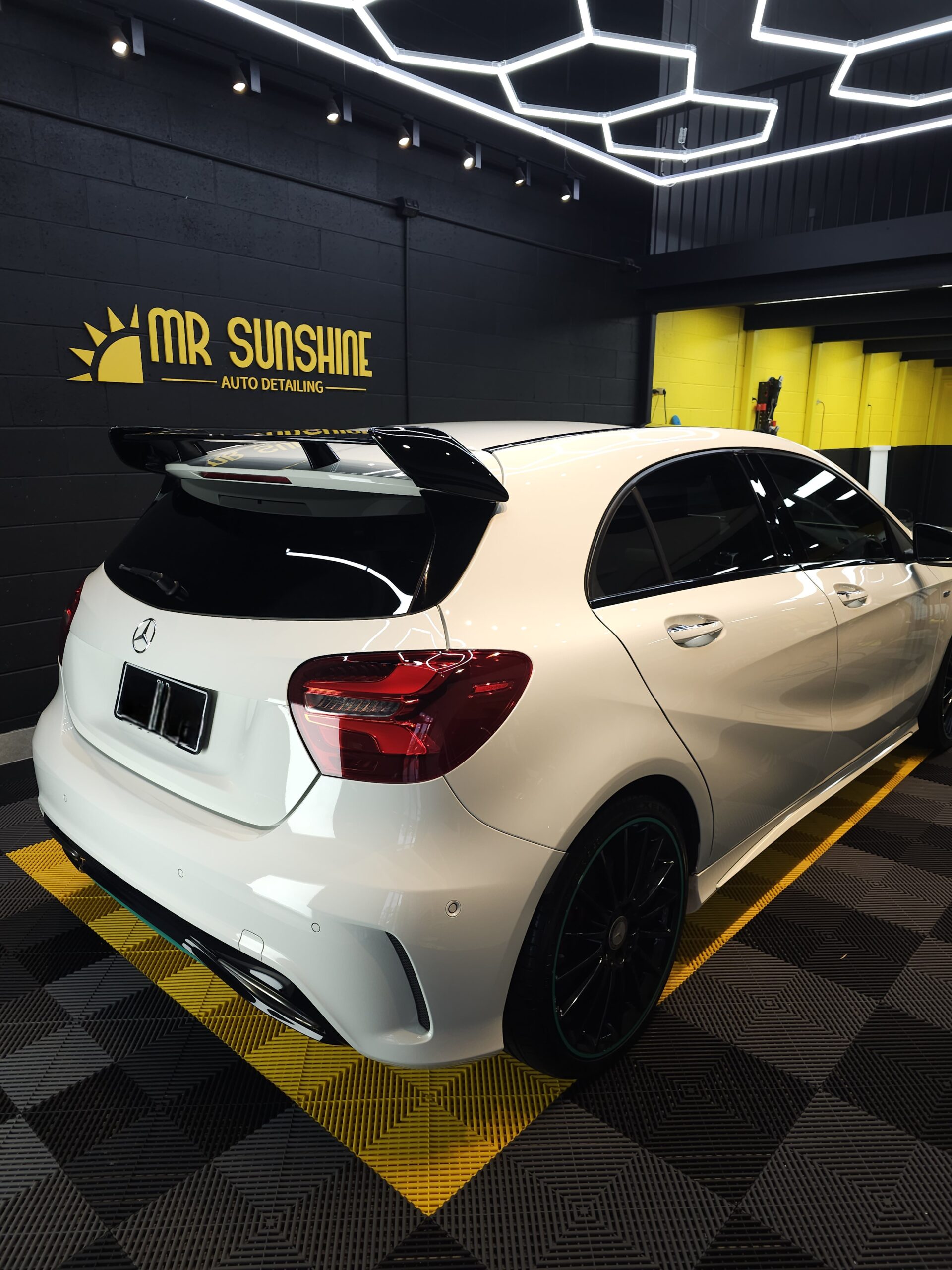 A white Mercedes-Benz hatchback with tinted windows and a rear spoiler is parked indoors at a car detailing shop named "Mr Sunshine Auto Detailing.