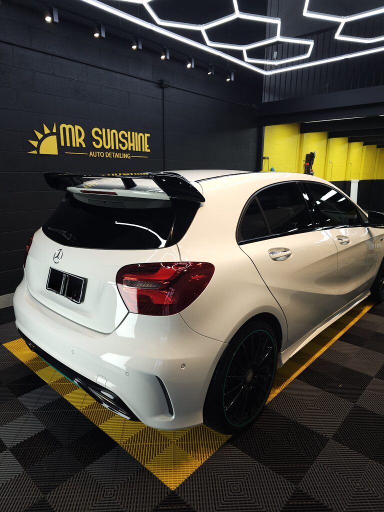 A white Mercedes-Benz hatchback is parked indoors at an auto detailing shop named "Mr Sunshine." Black and yellow accents are visible in the background. | Featured image of the Car Graffiti Removal page